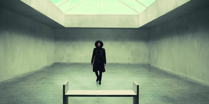 Colin Kaepernick, in a dark and sophisticated outfit, stands in the center of a light grey cement room under a large skylight. A minimalistic bench is in the foreground before him.